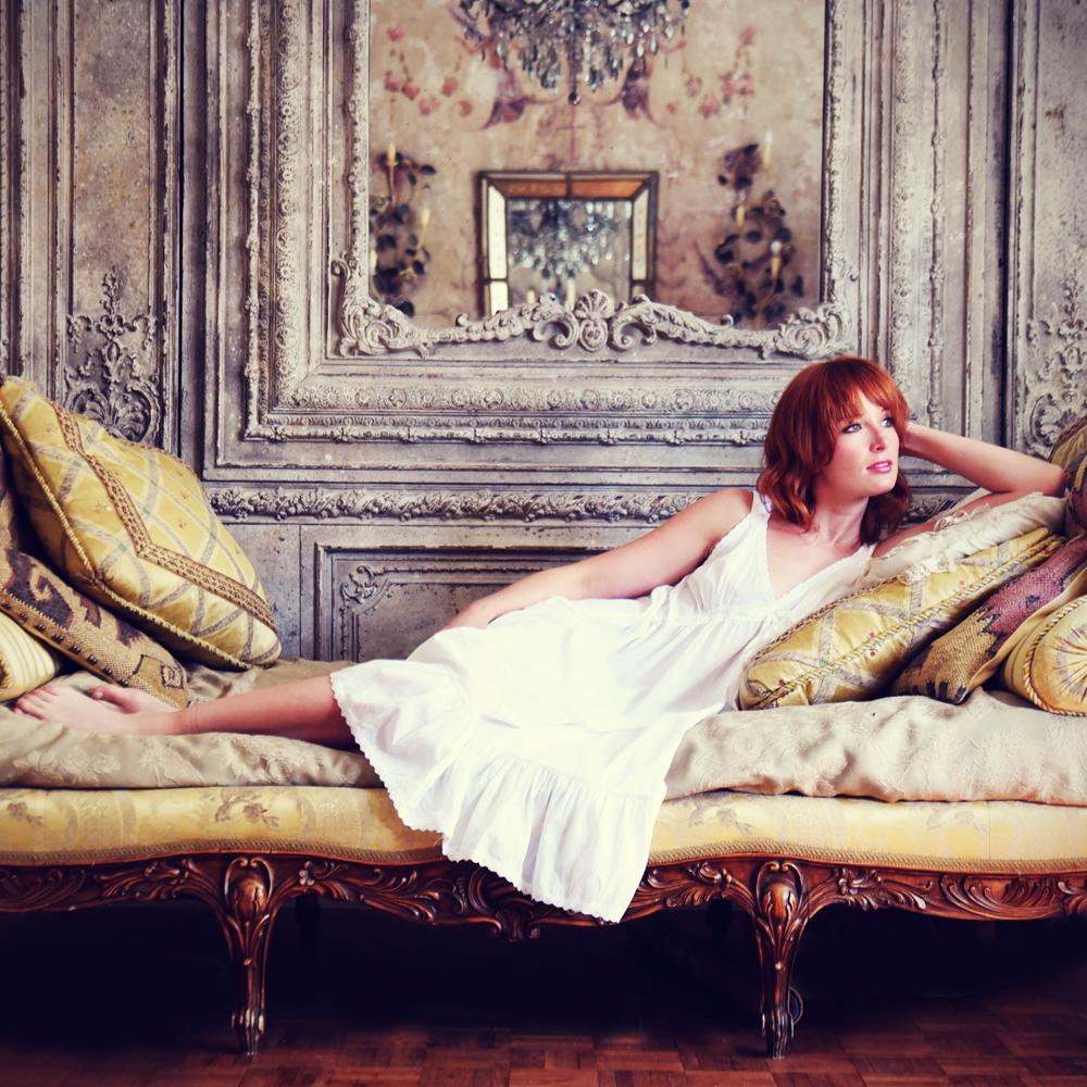 Red headed woman in white cotton nightie reclining on a yellow couch with cushions and an old fashioned background